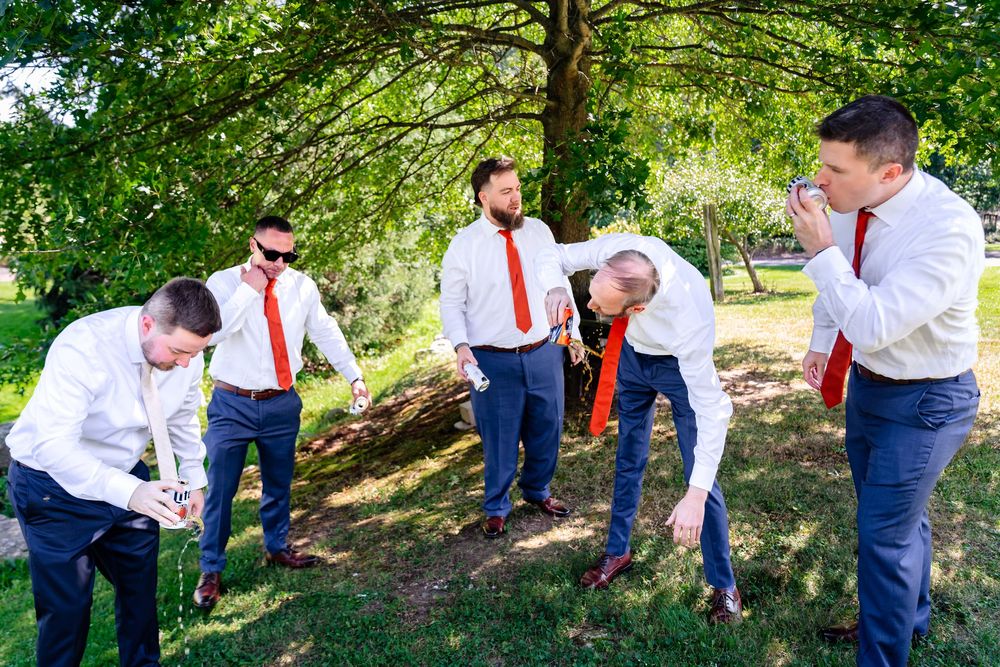 Groomsmen in white shirts and red ties adjusting neckwear outdoors under green trees before a wedding ceremony.