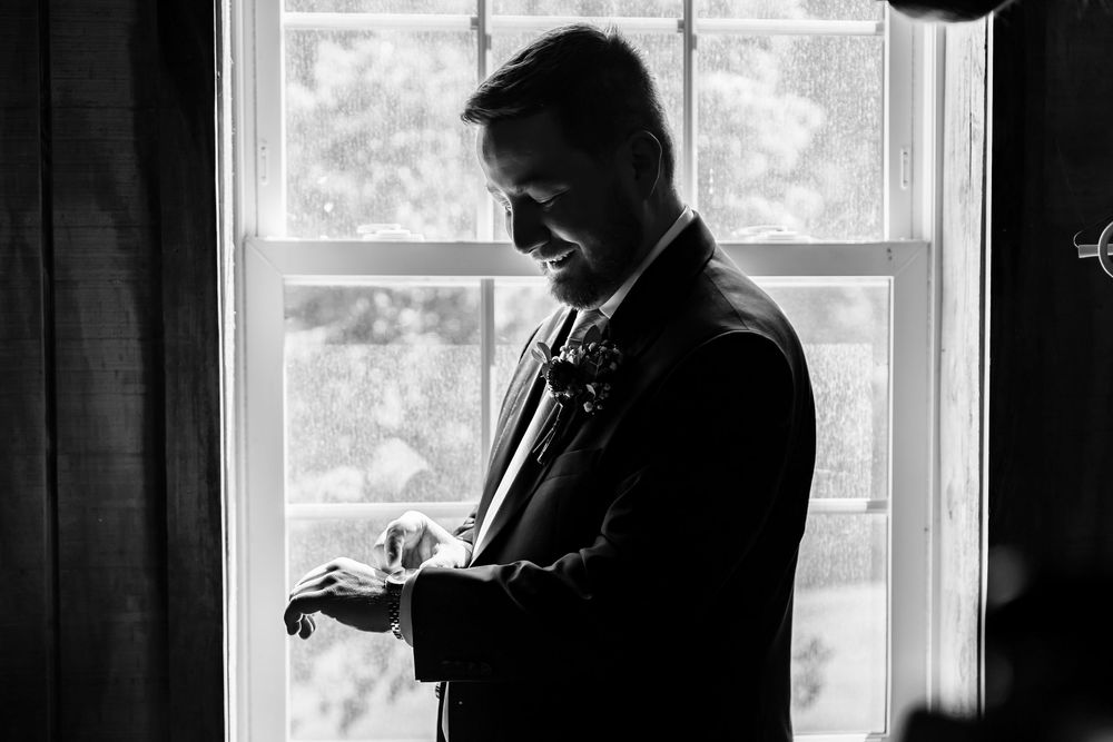 Groom in black suit adjusting boutonniere by window in elegant black and white wedding photo.
