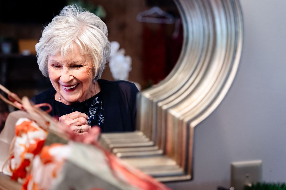 Elderly woman with white hair smiling joyfully while opening a gift wrapped in red ribbon at a celebration.