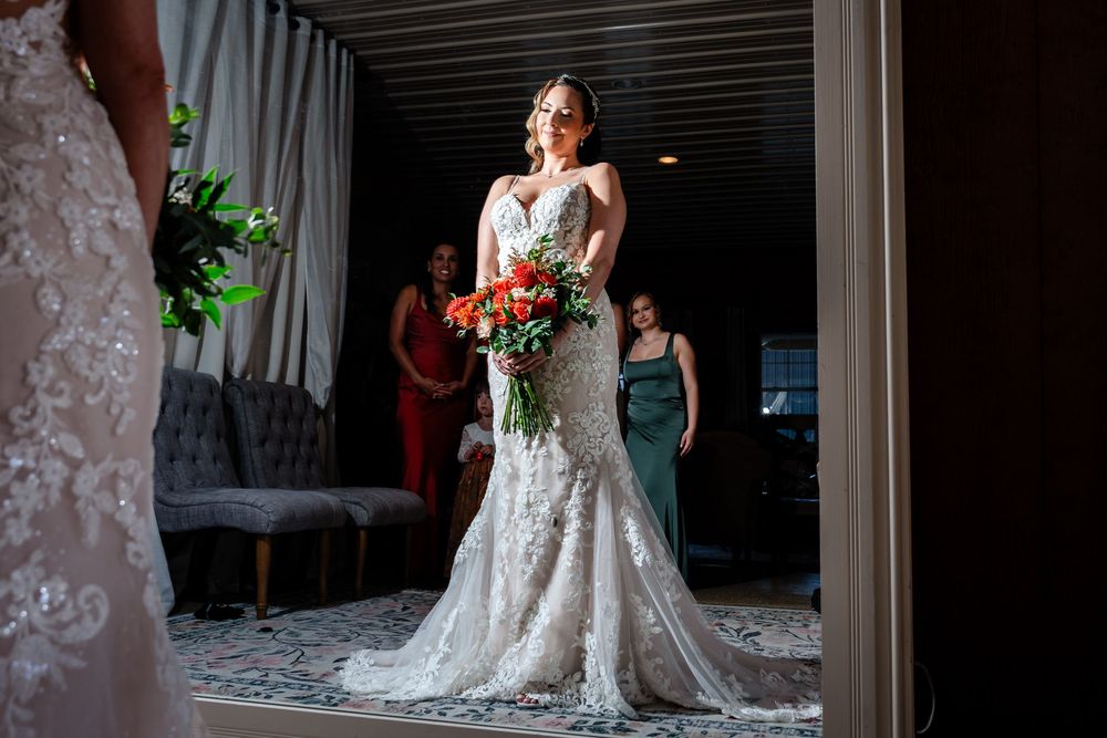 Bride in lace mermaid gown holds vibrant bouquet, bridesmaids in red and green dresses visible in background.