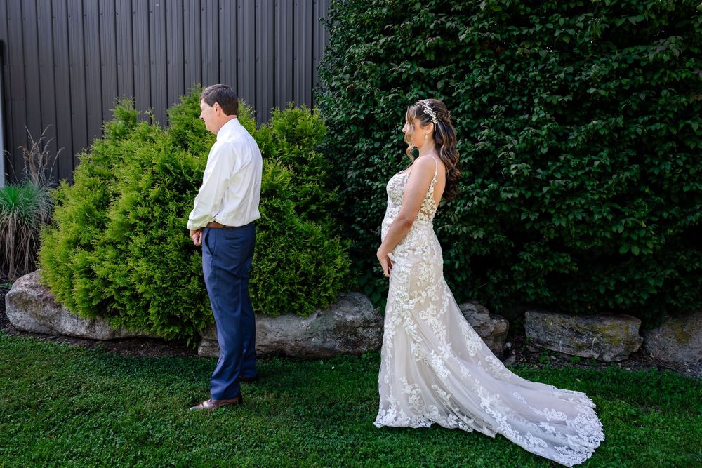 Bride in lace wedding gown and groom in white shirt share a first look moment on green lawn beside evergreen shrubs.