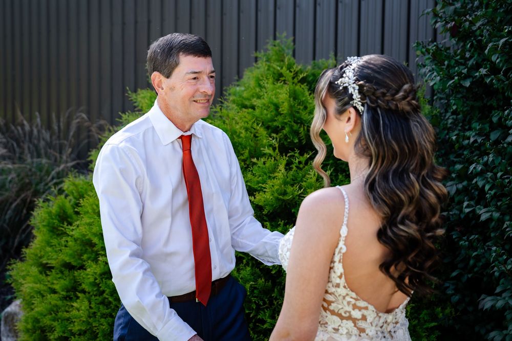 A man in a white shirt and red tie shares a joyful moment with a bride in a floral wedding dress outdoors.