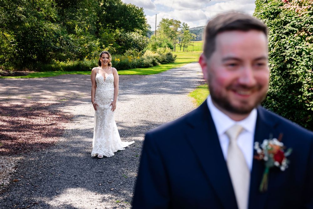 Groom in navy suit smiles in foreground as bride in lace wedding dress stands on gravel path surrounded by lush greenery.