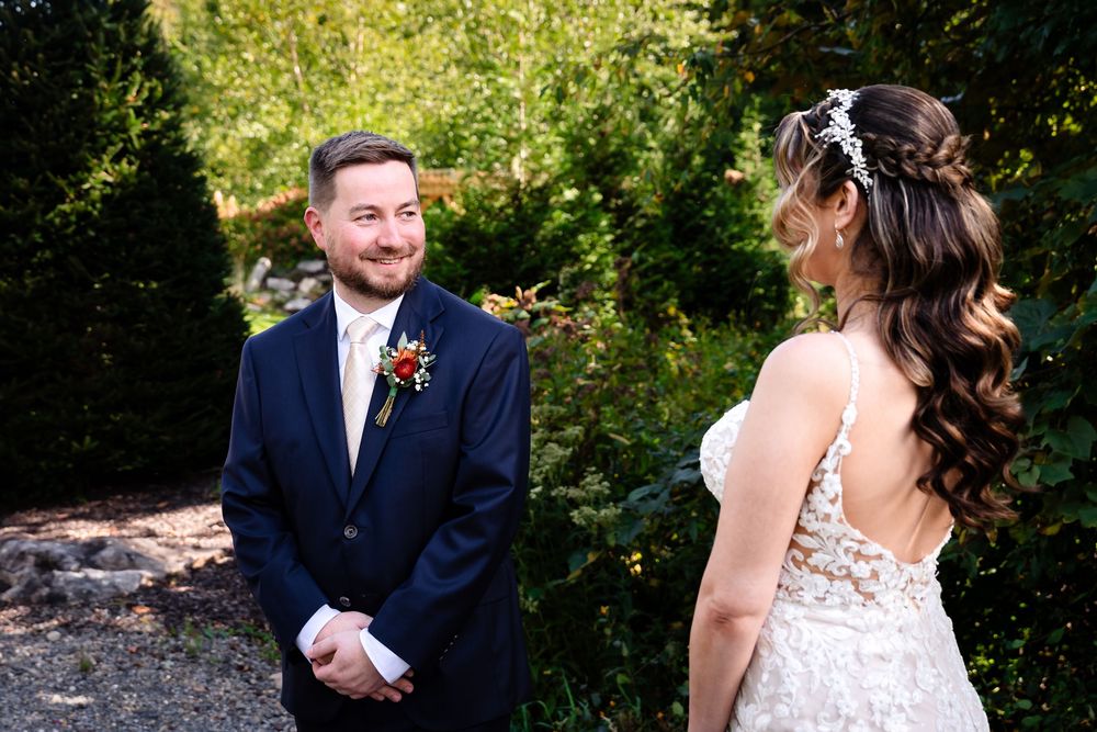 Groom in navy suit smiles at bride in lace wedding dress during outdoor first look ceremony.