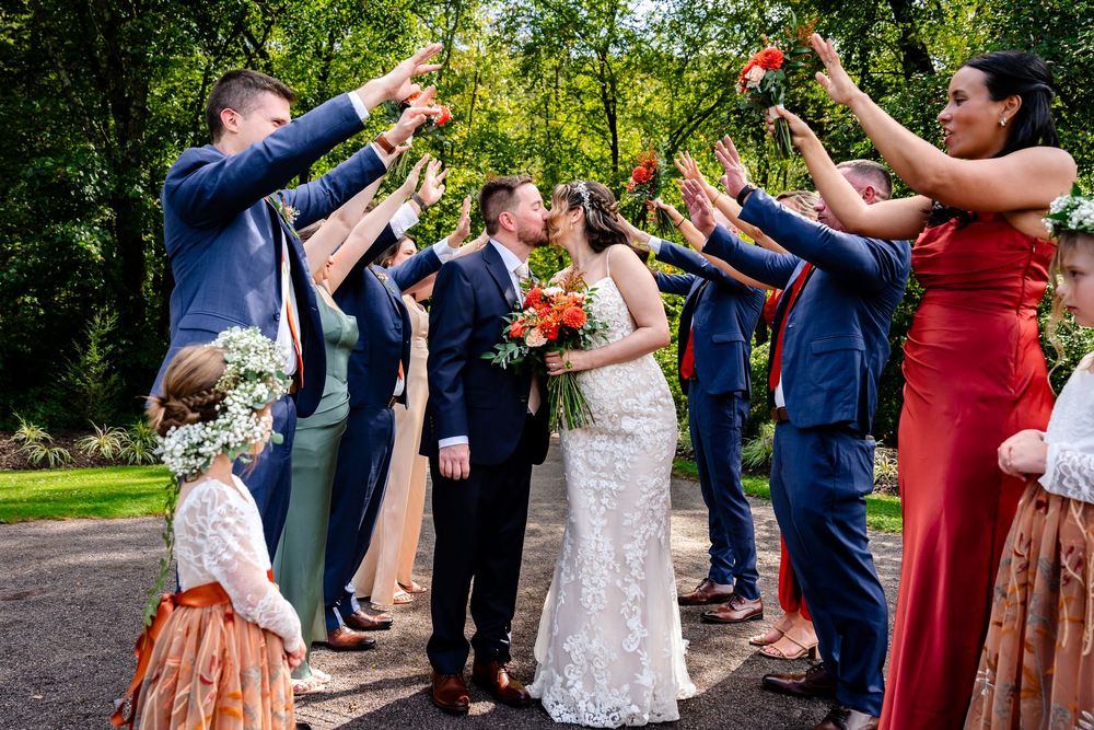 Wedding party cheering as bride and groom kiss under raised hands outdoors.
