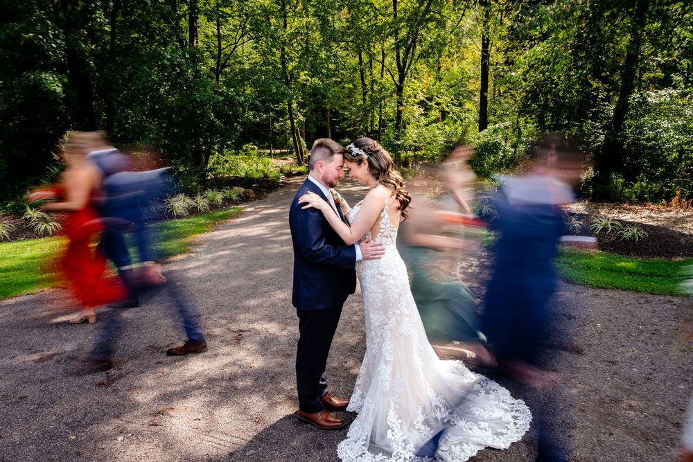 Bride and groom share a romantic moment as wedding guests blur past them on a sunlit garden path.