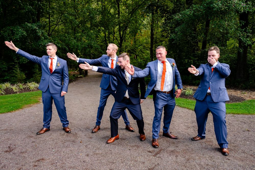 Five groomsmen in navy blue suits with orange ties pose playfully on a garden path surrounded by lush green trees.