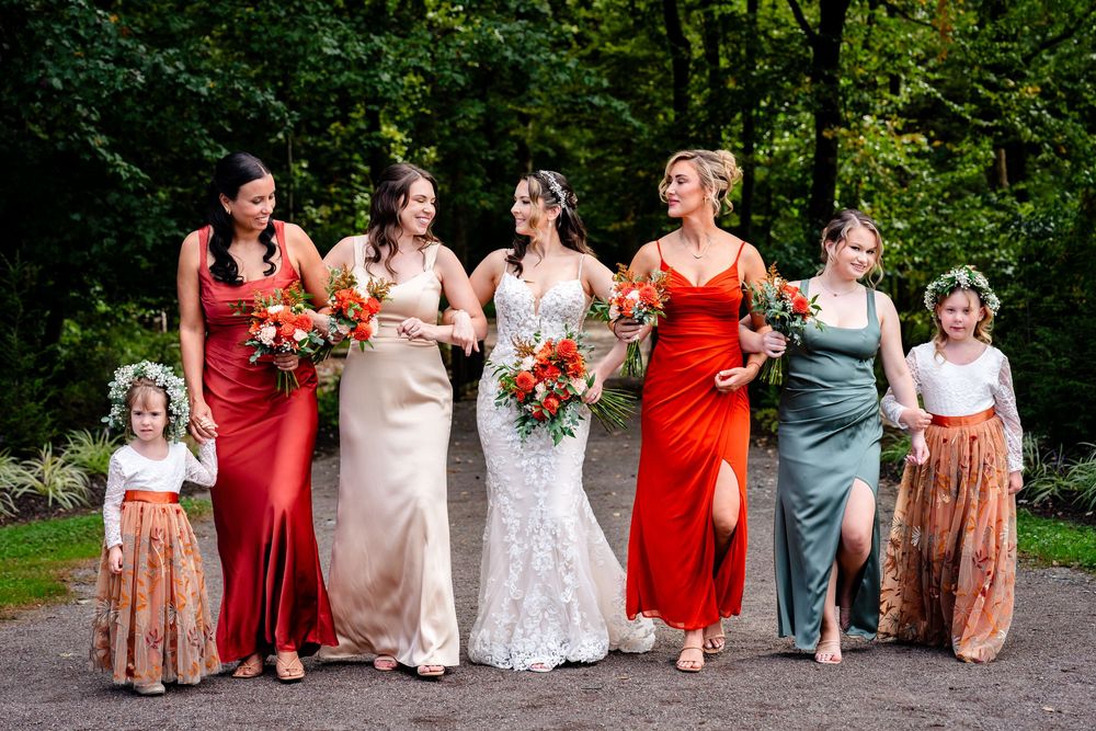 Bride with bridesmaids in red and sage gowns holding bouquets with flower girls outdoors.
