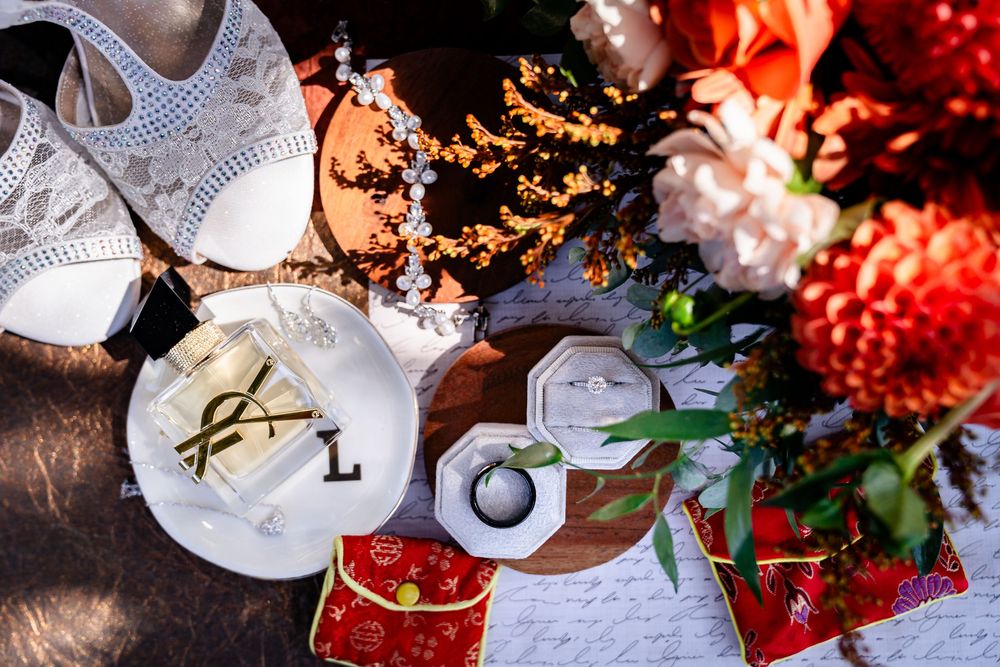 Wedding flat lay with bridal heels, rings, red floral bouquet, and decorative accessories on a wooden surface.