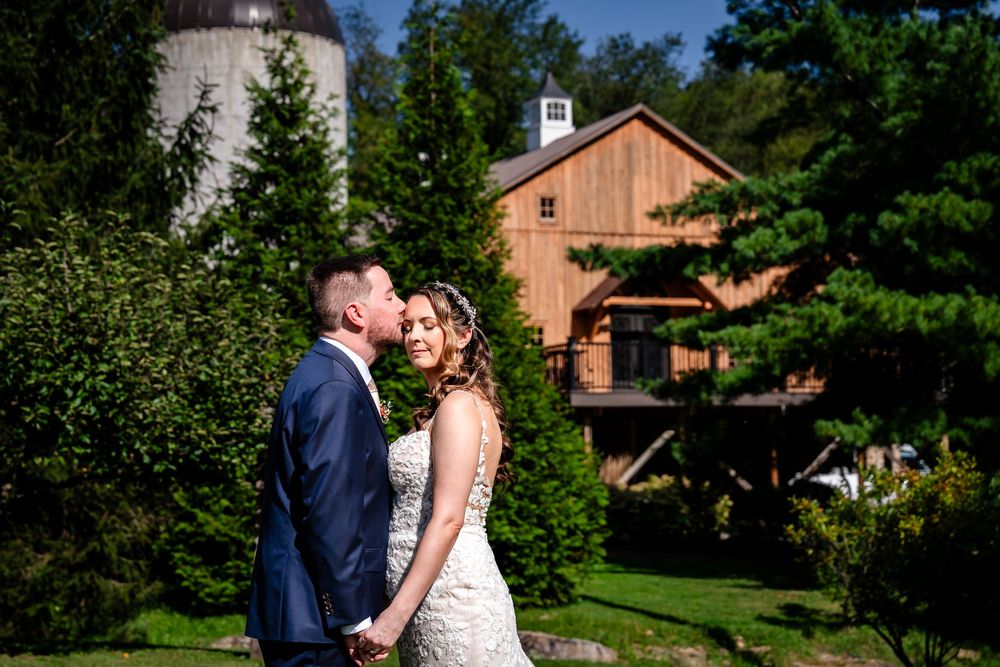 Bride and groom share a kiss outdoors with a rustic barn and silo backdrop on their wedding day.
