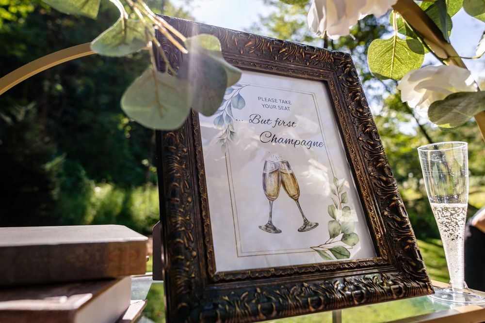 Elegant framed champagne toast sign at an outdoor wedding reception with string lights and greenery.