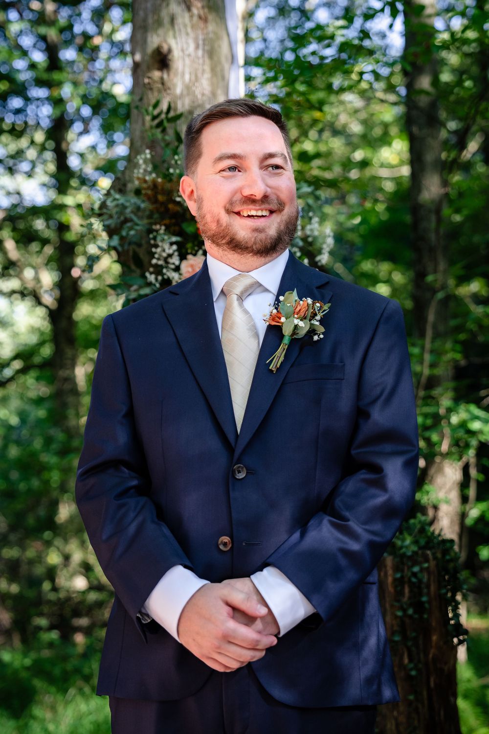 Smiling groom in navy suit with floral boutonniere stands outdoors among lush green trees.