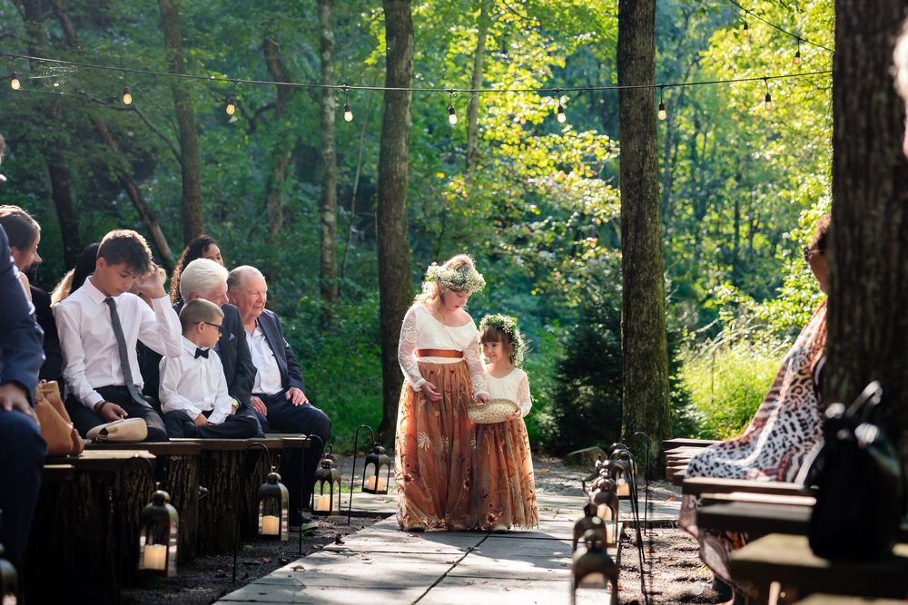 Bride walks down wooded outdoor aisle with flower girl during rustic forest wedding ceremony.