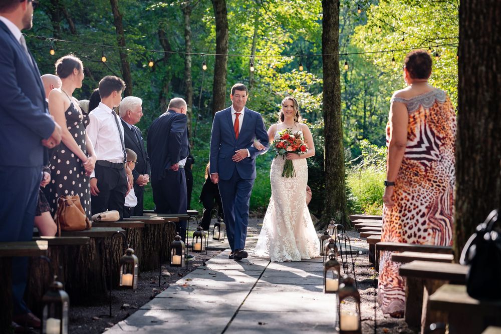 Bride walks down outdoor aisle with escort at woodland wedding ceremony, flanked by lanterns and guests.