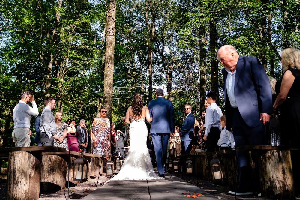 Bride walks down wooded outdoor aisle with guests seated on rustic benches at a forest wedding ceremony.