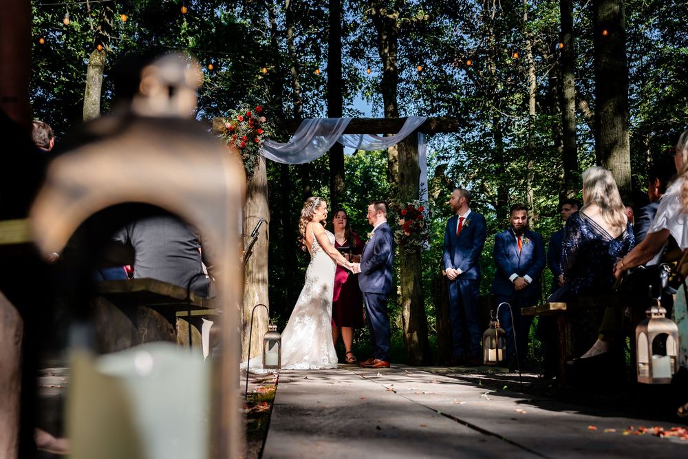Outdoor woodland wedding ceremony with guests seated along lantern-lined aisle.