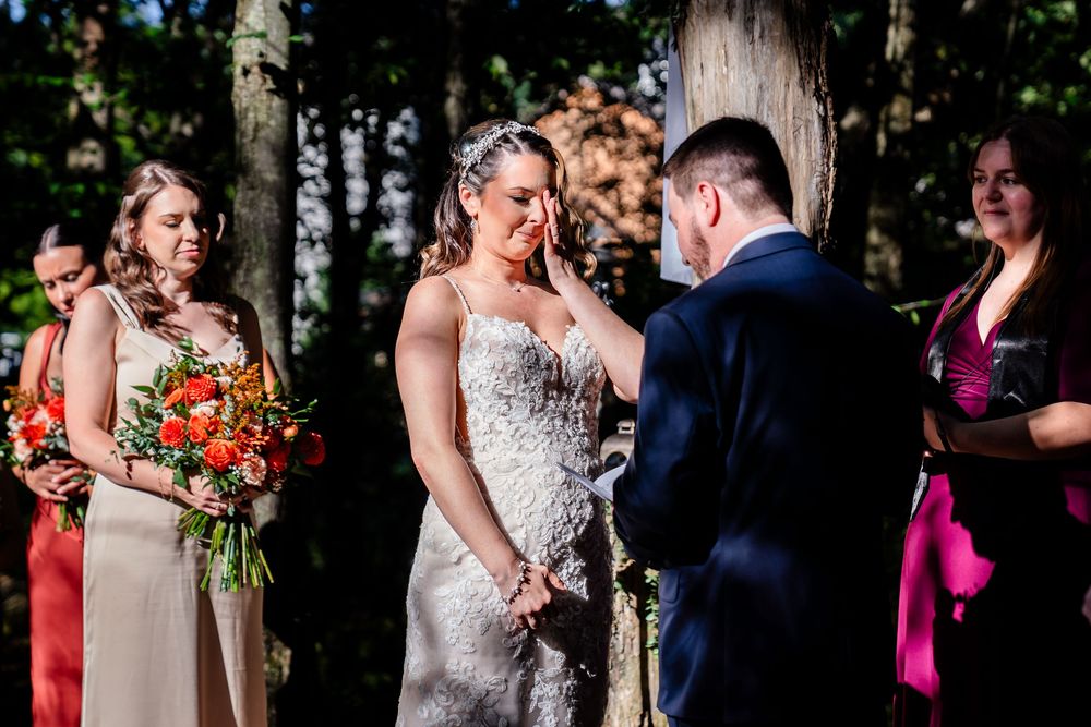 Groom gently wipes tears from brides face during an emotional outdoor wedding ceremony in a wooded setting.