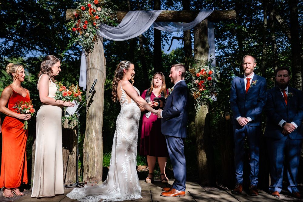 Couple exchanges vows under floral arch at outdoor woodland wedding with bridal party in orange and navy.
