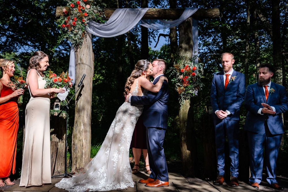 Bride and groom share first kiss under floral arch with wedding party at outdoor woodland ceremony.