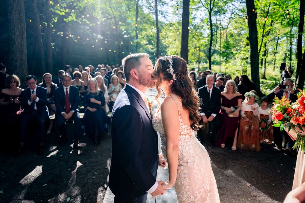 Bride and groom share first kiss at outdoor wedding ceremony surrounded by guests in a sunlit forest setting.