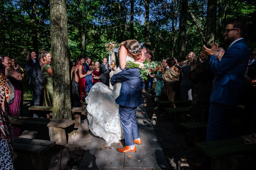 Groom lifts bride joyfully as guests celebrate their recessional at an outdoor forest wedding.