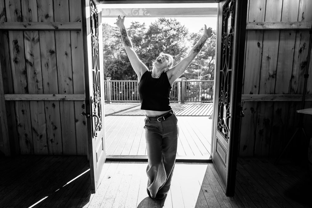 Black and white photo of a woman raising her arms in joy at open barn doors leading to a sunny outdoor deck.
