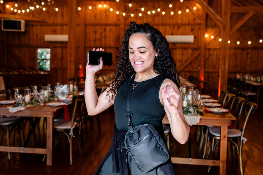 A smiling woman in a black dress holds a smartphone while dancing in a rustic barn venue with string lights.