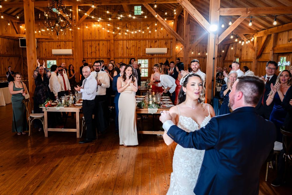 Bride and groom share first dance in warmly lit barn wedding venue as guests applaud.