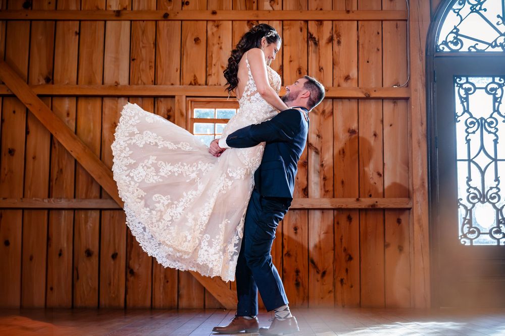 Groom lifts bride in lace wedding gown inside wooden barn with stained glass window.