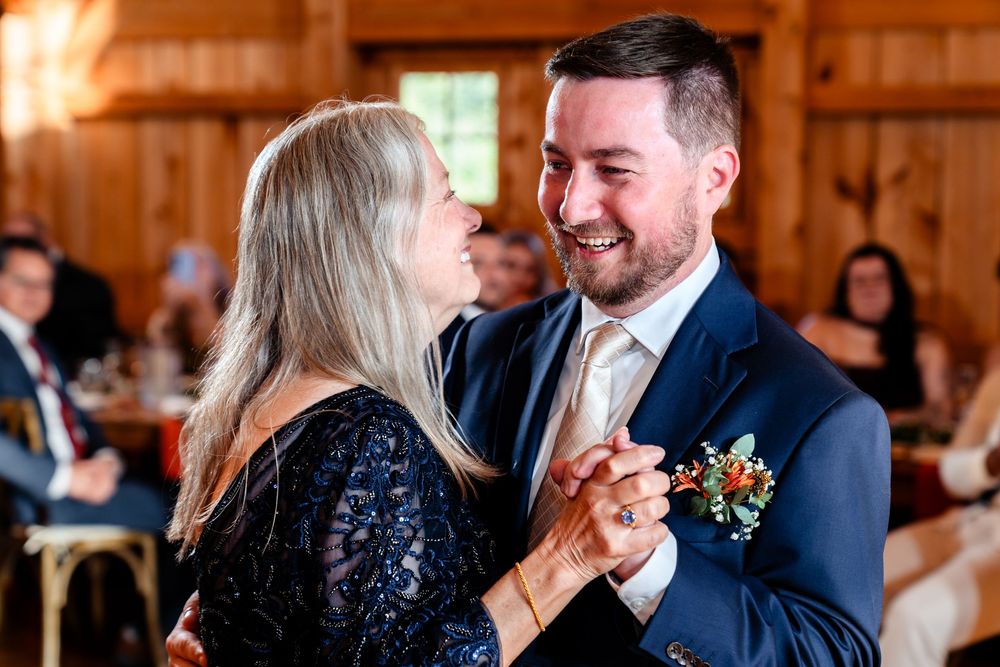 Groom shares a joyful mother-son dance in a rustic barn wedding reception venue.