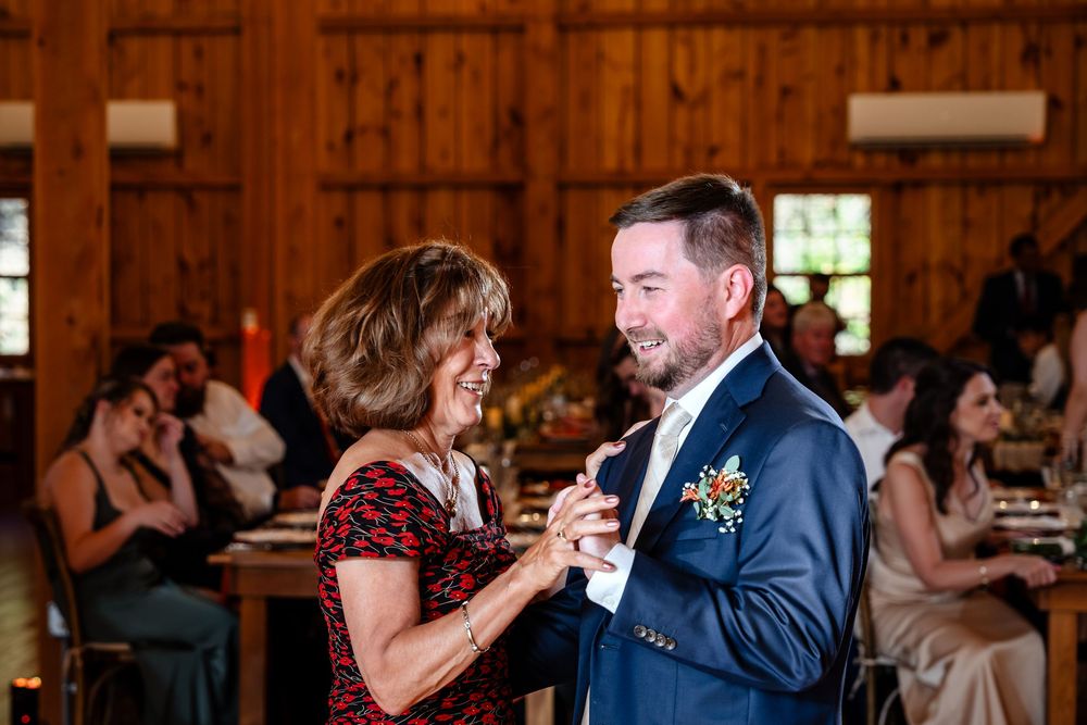 Groom dances with his mother during wedding reception in a warmly lit rustic barn venue.
