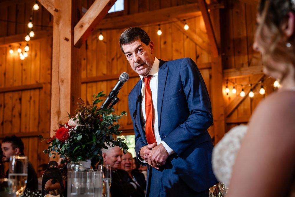 Man in blue suit with orange tie gives wedding speech at microphone in barn.