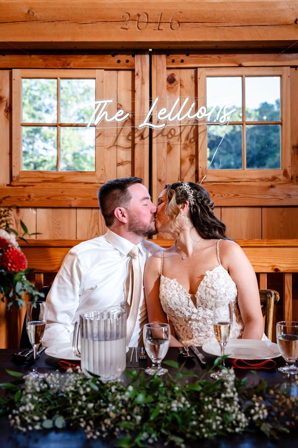 Newlyweds sharing a kiss at their rustic barn wedding reception sweetheart table with champagne glasses and floral centerpieces.
