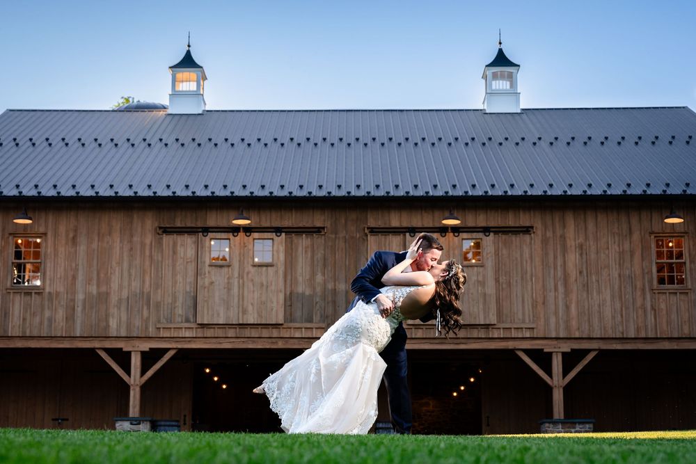 Groom dips bride in front of rustic barn venue under clear blue sky.