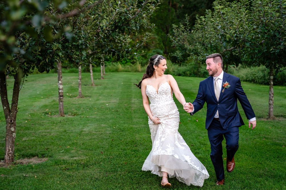 Bride and groom walk hand in hand through lush green orchard in wedding attire.