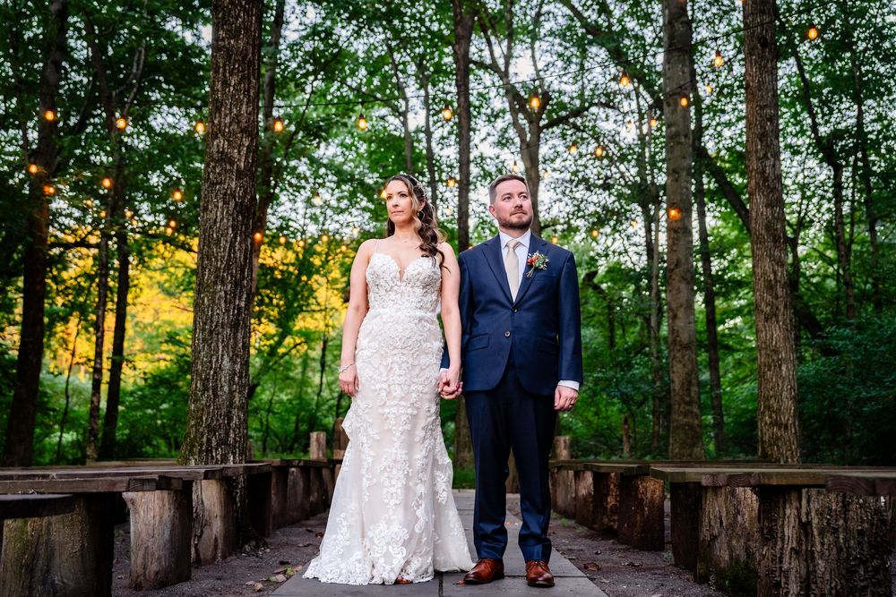 Bride and groom holding hands on a wooden bridge surrounded by tall trees with string lights at golden hour.