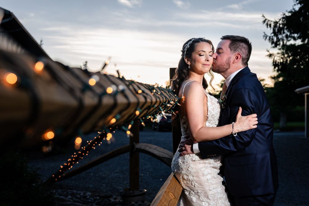 Bride and groom share a romantic kiss at sunset beside a wooden fence adorned with warm string lights.