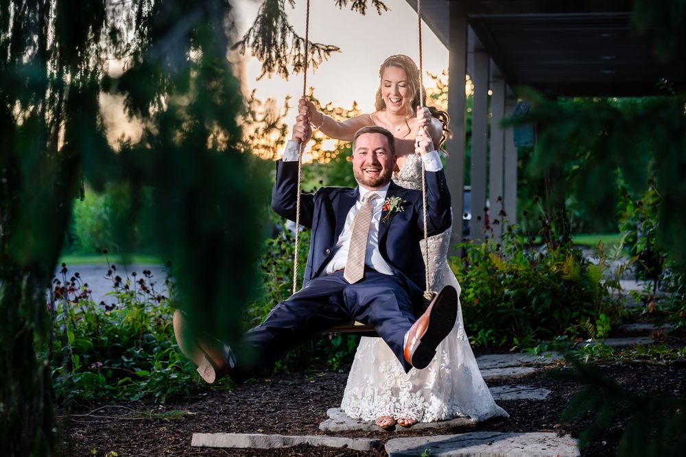 Joyful bride and groom play on a wooden swing surrounded by a lush garden at golden hour.