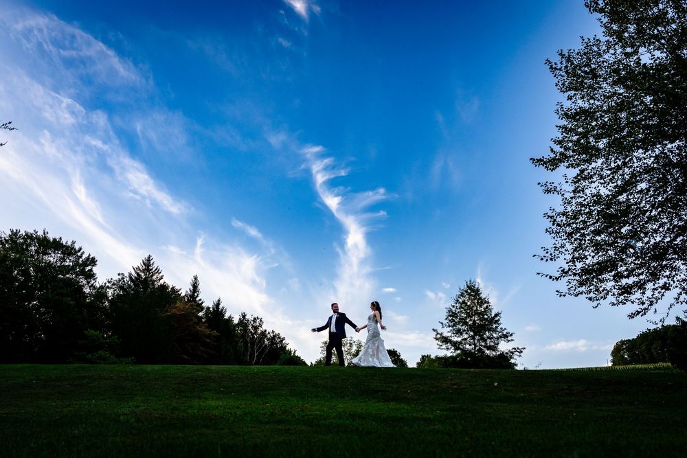 Bride and groom walk hand in hand across a green hill under a dramatic blue sky on their wedding day.