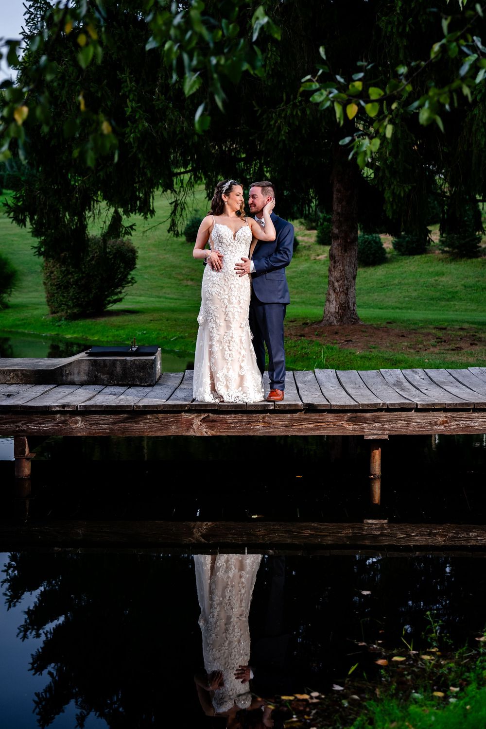 Bride and groom embrace on a wooden dock reflected in still water beneath a lush green tree.