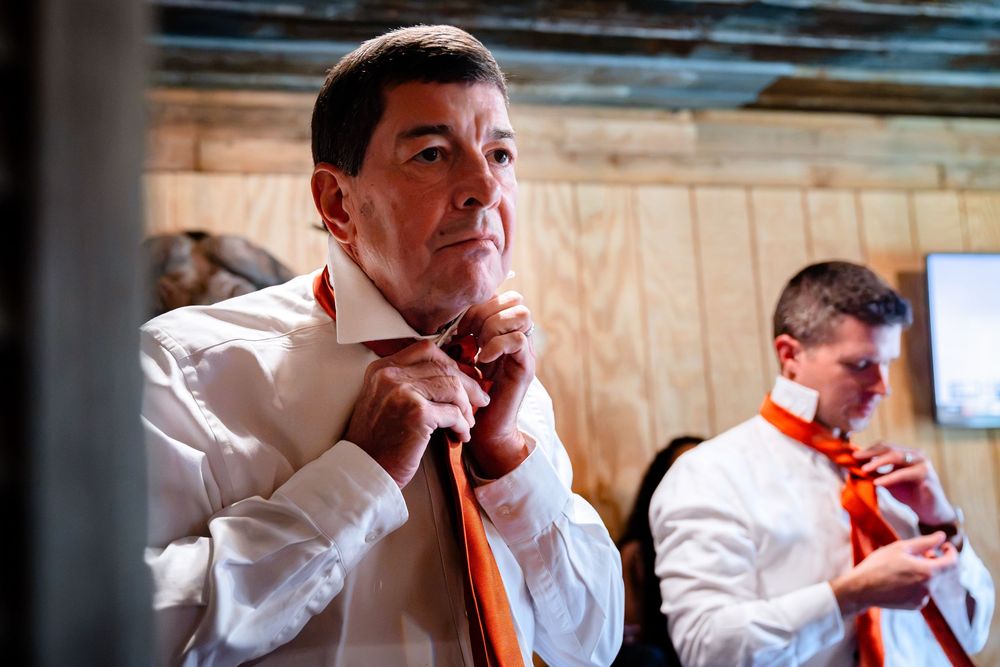 Two men in white shirts adjusting orange ties, preparing for a formal event indoors.