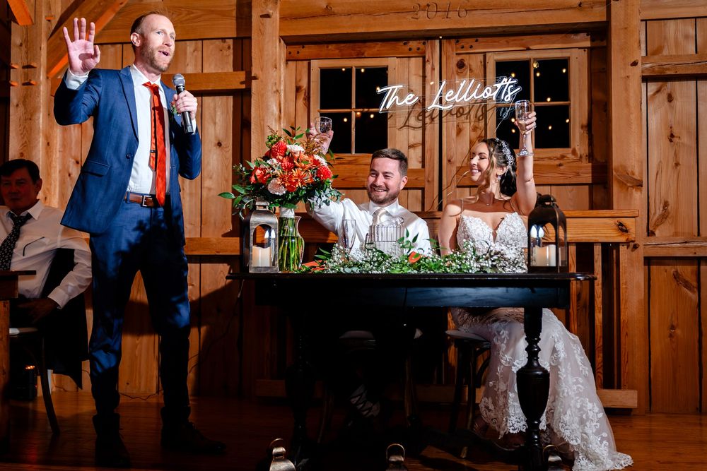 Wedding guests laugh during a heartfelt speech inside a rustic barn reception venue with warm lighting.