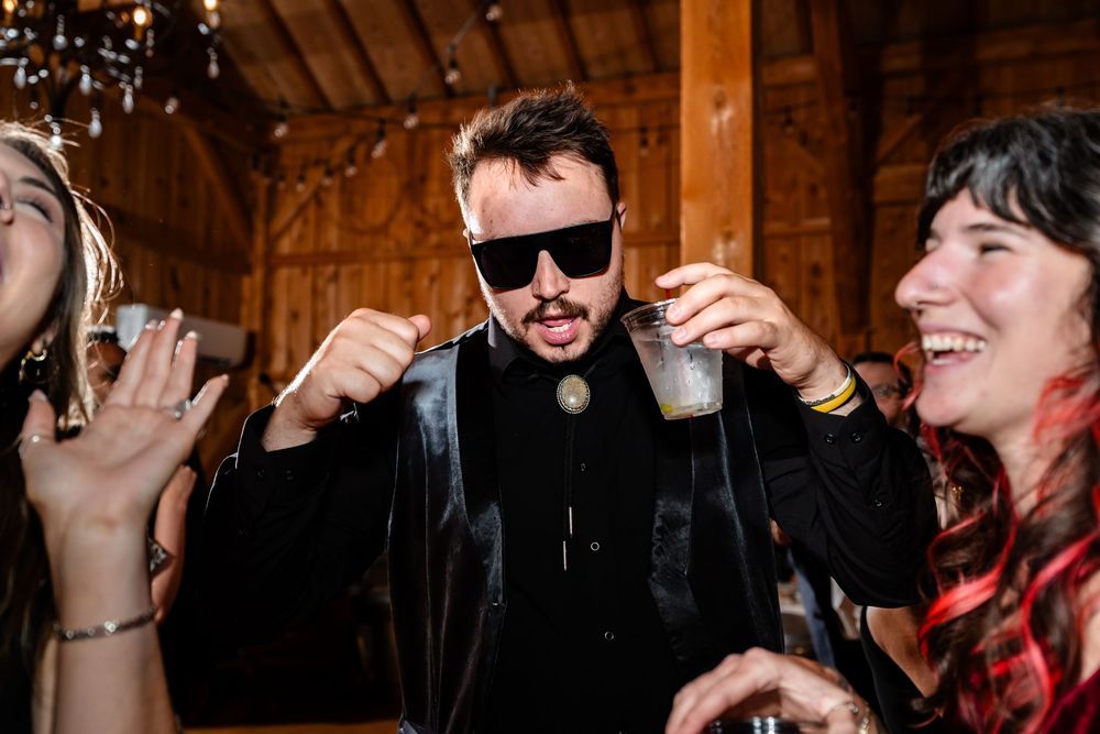 Guest in black suit and sunglasses celebrates at lively barn wedding reception.