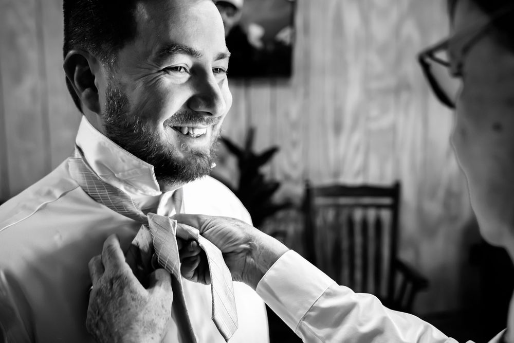Black and white photo of a smiling groom having his tie adjusted by someone at a wedding preparation.