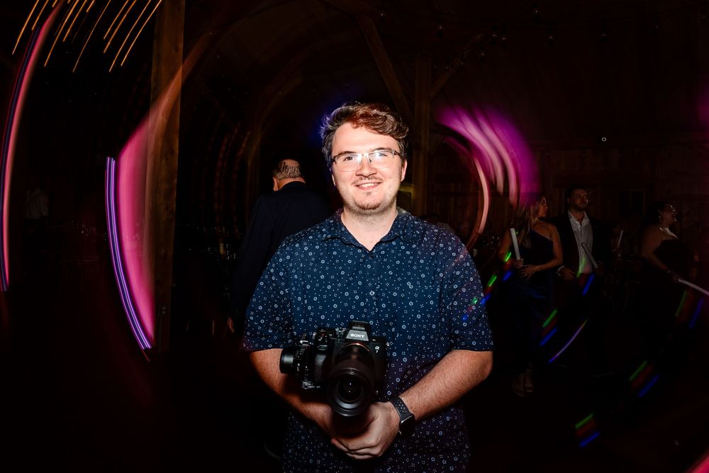 Wedding photographer smiles while holding a camera at a colorfully lit wedding reception venue.