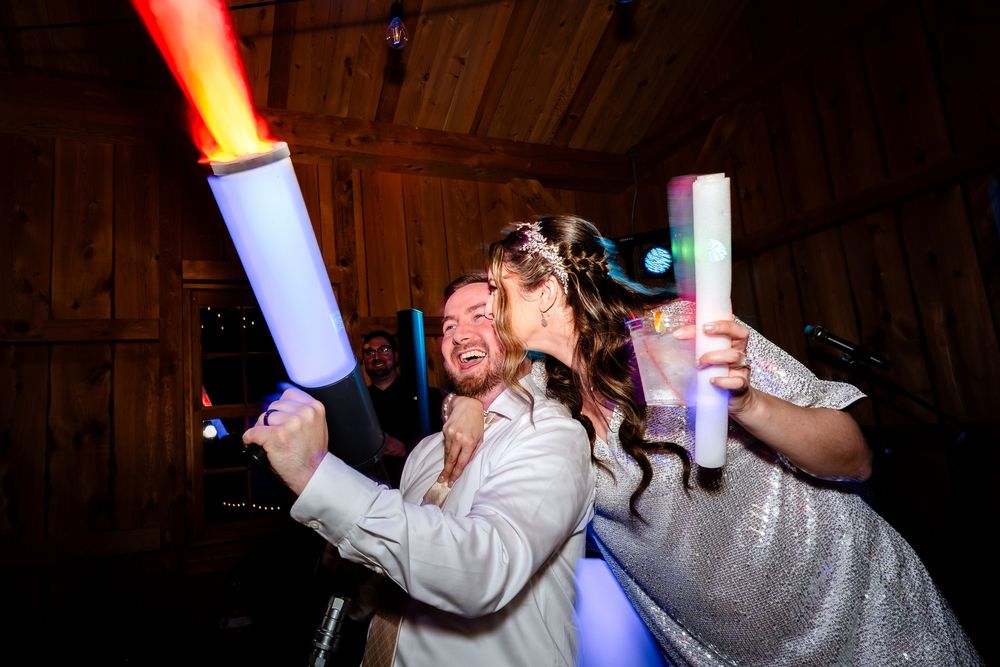 Bride and groom celebrate at wedding reception holding colorful LED foam sticks in a rustic barn venue.