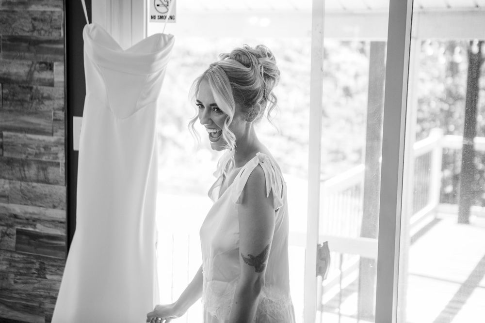 Black and white photo of a smiling bride in a slip dress standing next to her hanging wedding gown near a window.