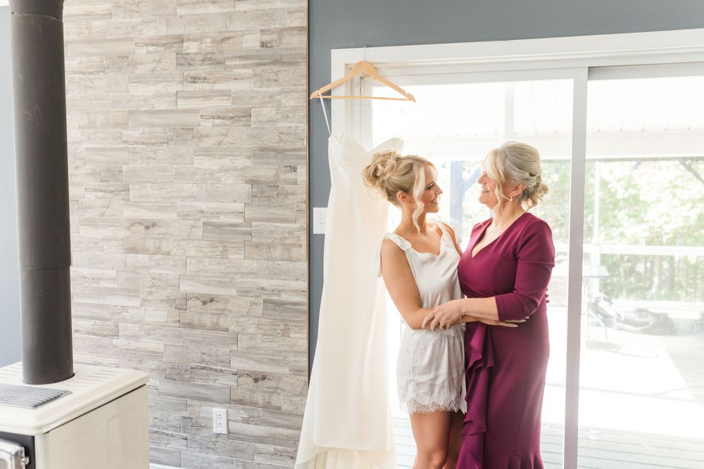 Bride and mother sharing a tender moment before wedding ceremony, with white gown hanging in background.