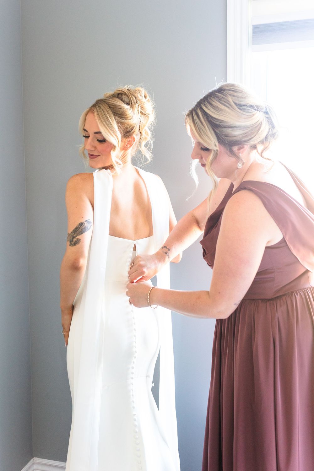 Bridesmaid in mauve dress fastening buttons on bride's white halter wedding gown before ceremony.