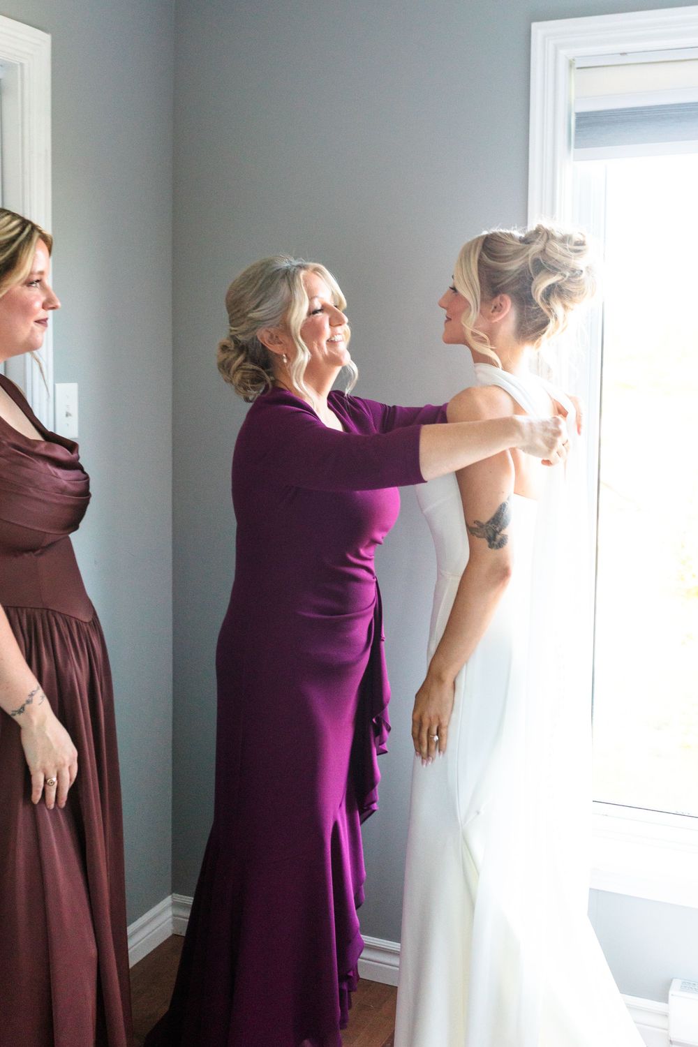 Mother in purple dress helps bride in white gown prepare, bridesmaid in burgundy looks on near window.